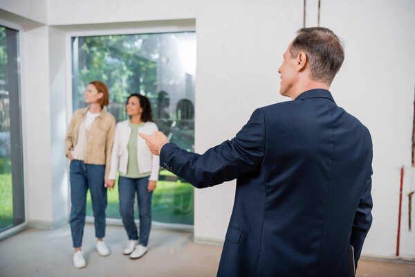 realtor pointing at panoramic window near blurred interracial lesbian couple inside of new house
