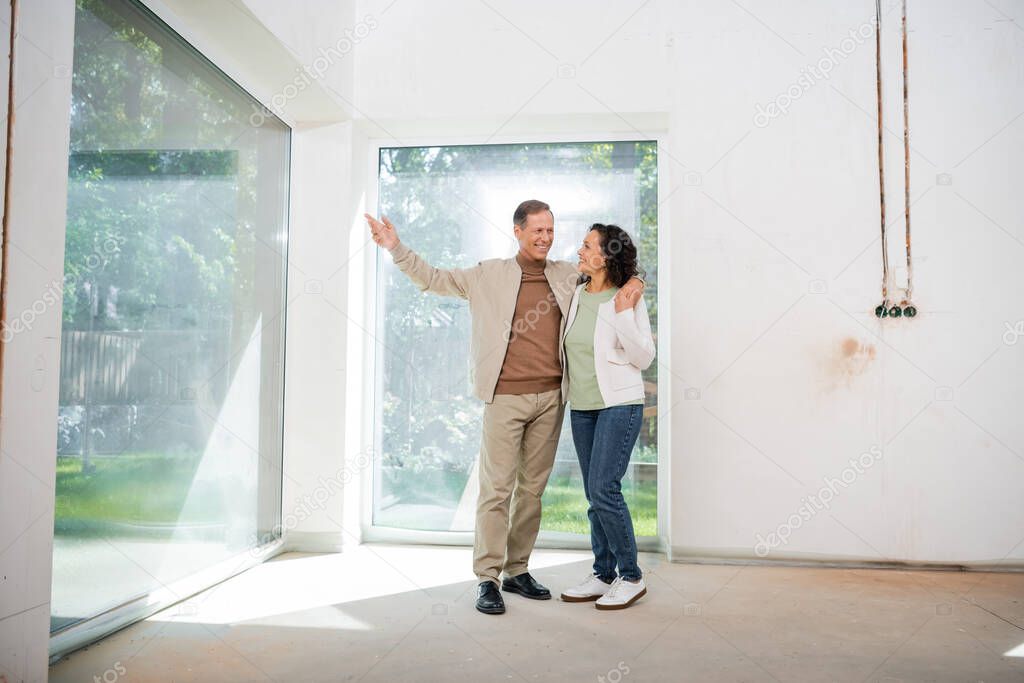 happy man pointing with hand at panoramic windows while hugging wife in new house