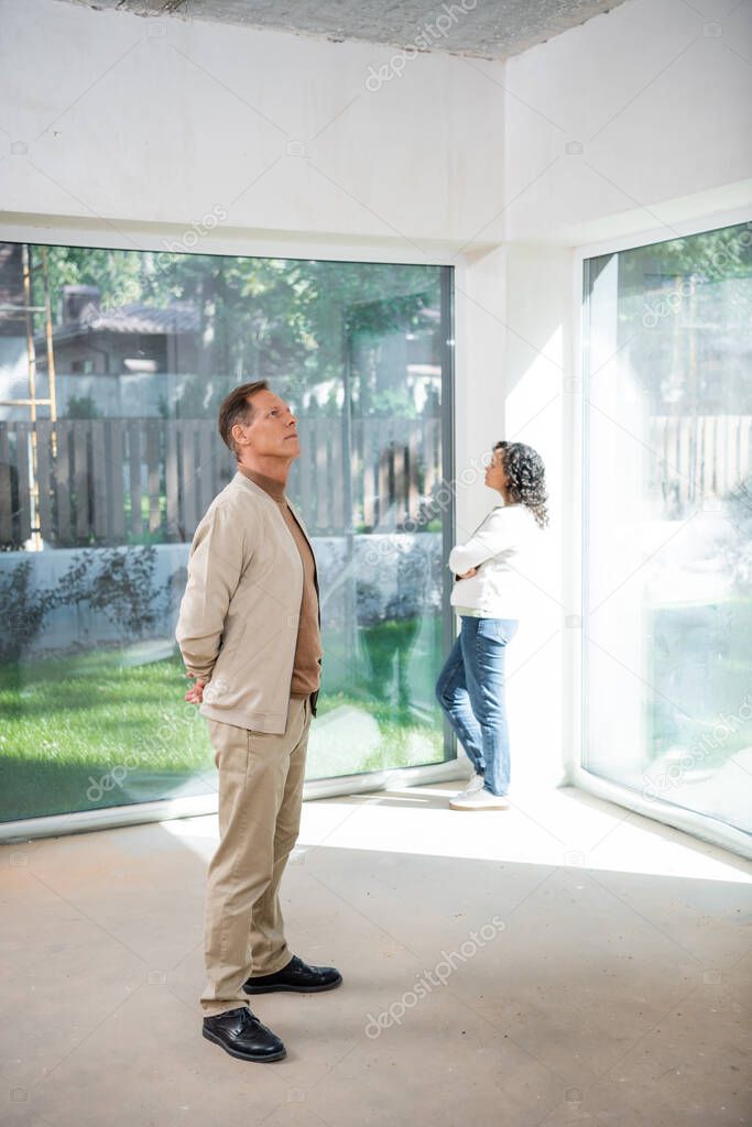 man looking at ceiling near african american wife standing near panoramic windows in new house