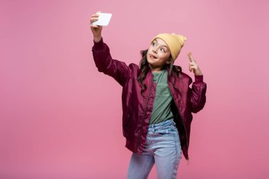 Şık giyinmiş genç kız selfie çekiyor ve pembe üzerinde izole edilmiş barış işareti gösteriyor. 