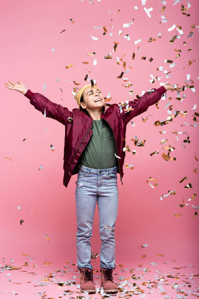 full length of joyful preteen girl in stylish outfit smiling near falling confetti on pink 