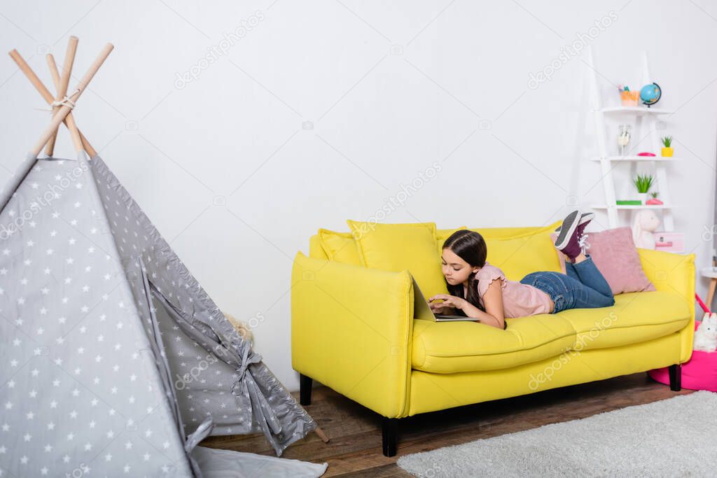 preteen girl lying on couch and using laptop in modern living room 