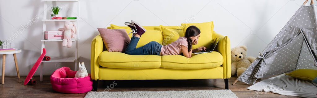 preteen girl lying on sofa and using laptop in modern living room, banner