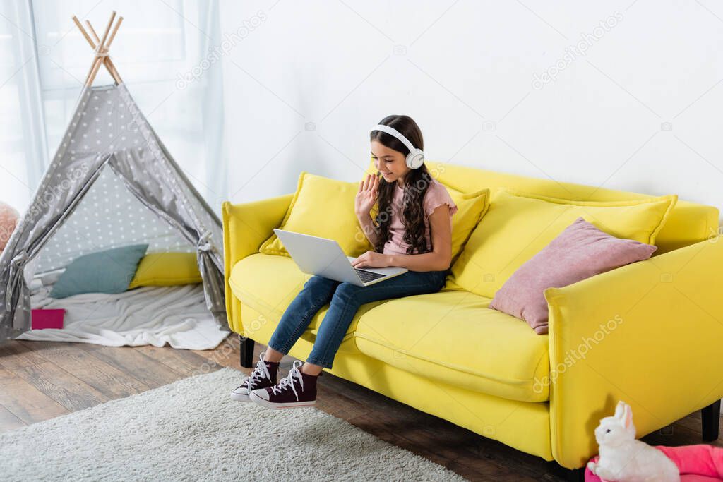 smiling preteen girl in wireless headphones waving hand during call while studying online at home