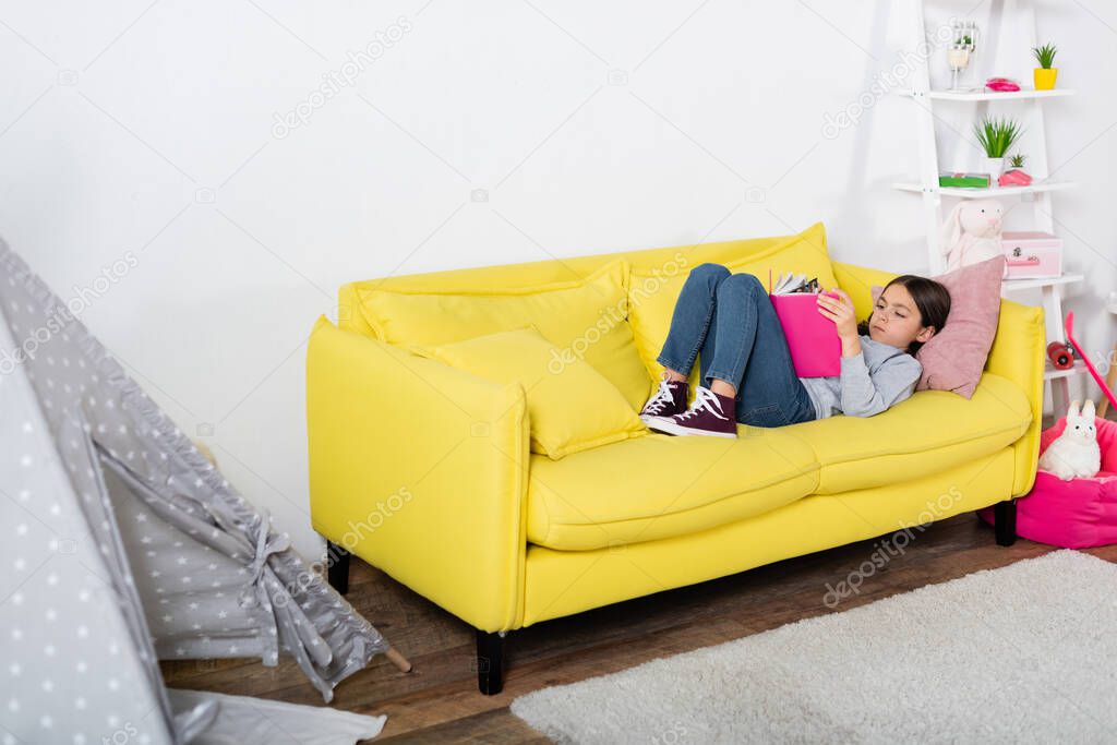 preteen child reading book while lying on couch at home