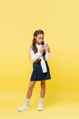 Sarı arka planda cep telefonu kullanan etekli okul çocuğu. 