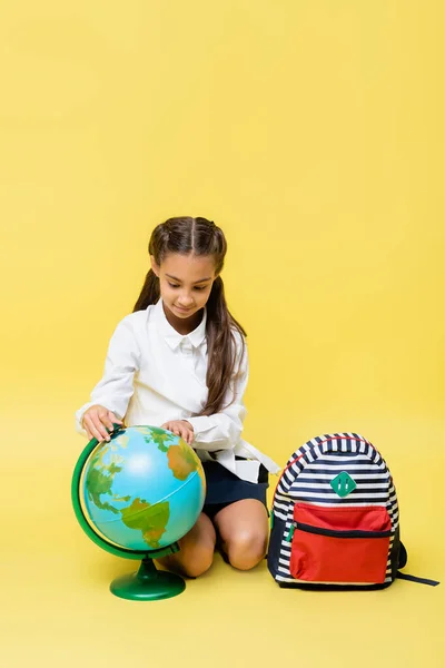 黄色の背景にバックパックの近くに地球を見て学校の子供 — ストック写真