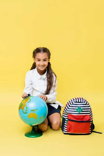 Szczęśliwe Dziecko Patrząc Aparat Pobliżu Globu Plecak Żółtym Tle — Zdjęcie stockowe