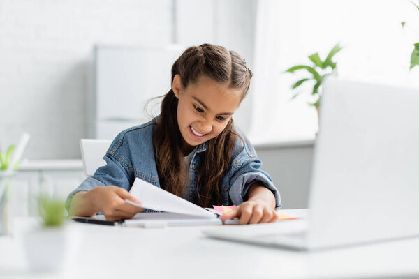 Angry preteen kid tearing paper from notebook near blurred laptop on table at home