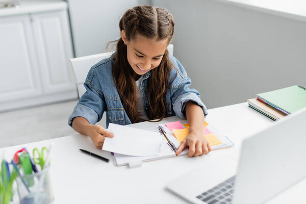 Happy kid tearing paper near notebook with sticky notes and blurred laptop at home
