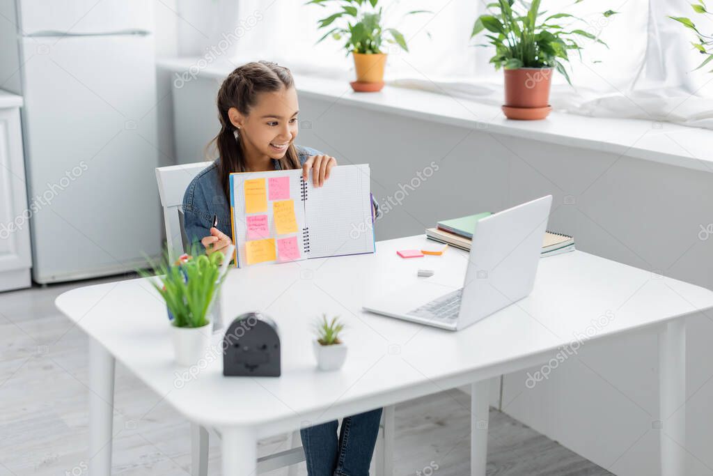 Smiling schoolgirl holding notebook with sticky notes near laptop during online education at home 