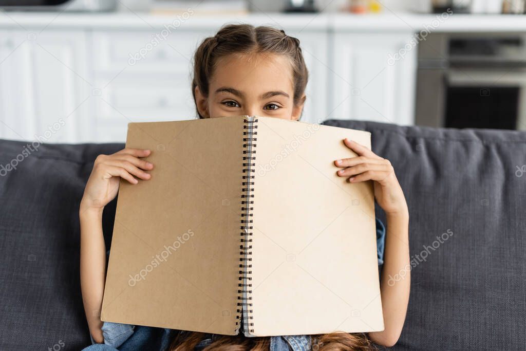 Preteen kid covering face with notebook and looking at camera on couch at home 