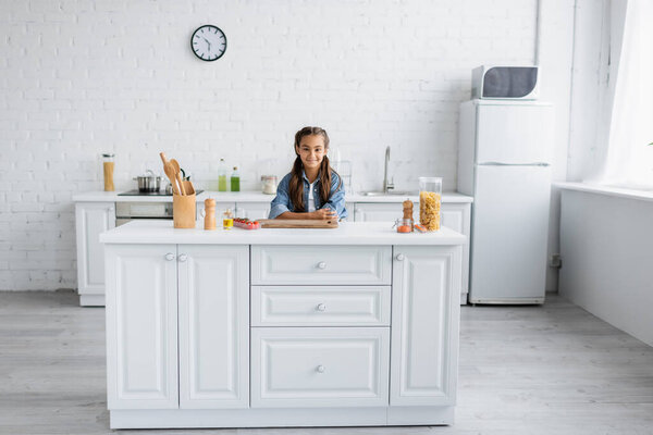 Smiling kid looking at camera near macaroni and cherry tomatoes in kitchen 