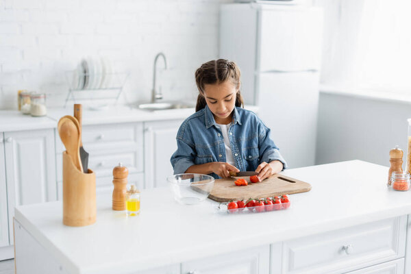 Preteen girl cutting cherry tomato near olive oil and bowl in kitchen 