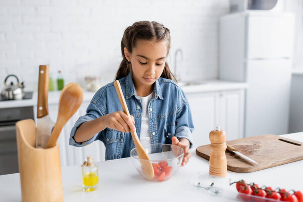Preteen kid mixing cherry tomatoes in bowl near cutting board and olive oil in kitchen 