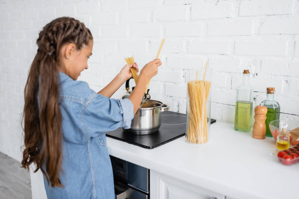Blurred kid holding macaroni near saucepan on stove at home 
