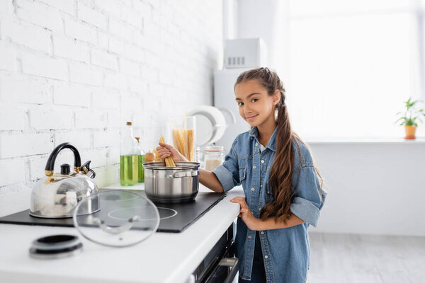 Smiling child holding macaroni near saucepan and looking at camera in kitchen 