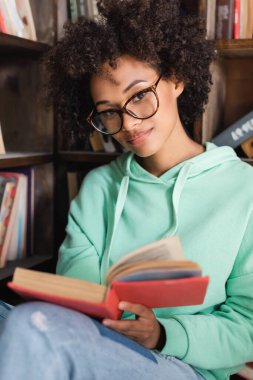 Gözlüklü genç Afrikalı Amerikalı öğrenci kütüphanede kitap tutuyor. 