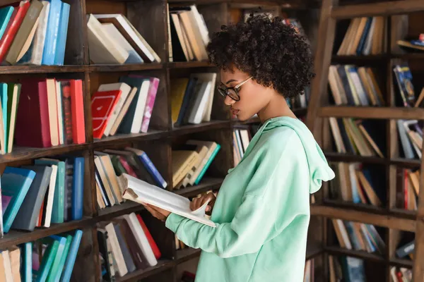 Молодая Африканская Американская Студентка Очках Читает Книгу Возле Книжного Шкафа — стоковое фото