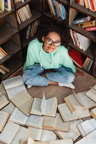 high angle view of happy african american student in eyeglasses sitting with crossed legs surrounded by books in library 