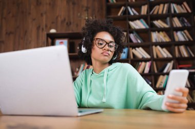 Bulanık dizüstü bilgisayarın yanında gözlüklü Afro-Amerikan öğrenci akıllı telefon kullanıyor. 