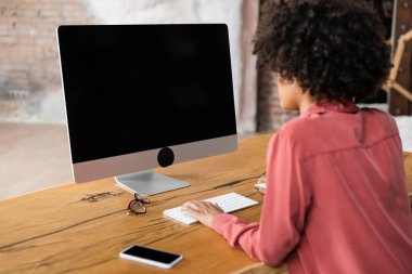 Kıvırcık Afrikalı Amerikalı kadın akıllı telefonun yanında bilgisayar klavyesi ve masasında gözlük kullanıyor.