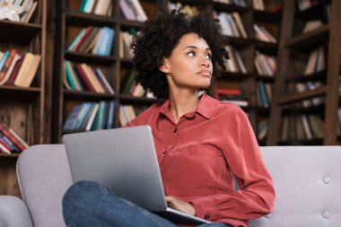 distracted african american woman sitting on couch with laptop and looking away clipart