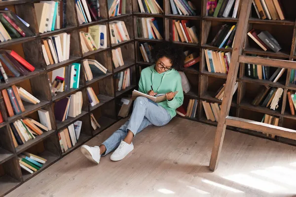 アフリカ系アメリカ人の若い学生が本に囲まれた床に座って図書館でノートを書き — ストック写真