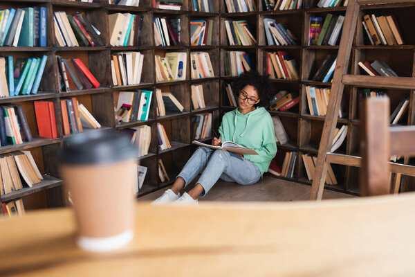 young african american student in eyeglasses sitting on floor and writing on notebook near blurred paper cup on foreground