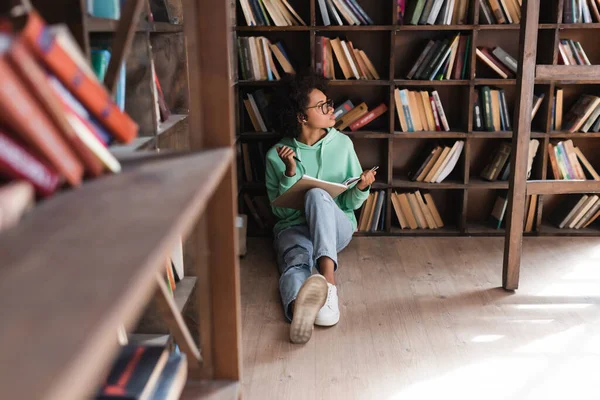 Estudiante Afroamericano Pensativo Gafas Sentado Sosteniendo Cuaderno Biblioteca Con Estantería — Foto de Stock