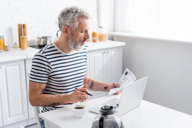 Elinde banknot ve kalemle elinde kağıt olan orta yaşlı bir adamın evde laptopun ve kahvenin yanında görüntüsü. 