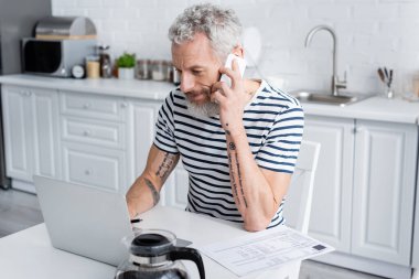 man talking on smartphone and using laptop near bills and coffee at home. Translation: 