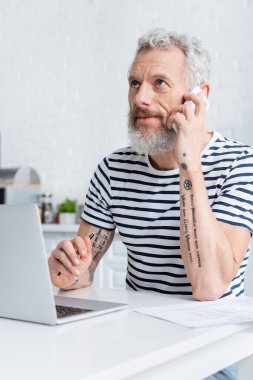 mature man talking on cellphone near papers and laptop in kitchen. Translation: 