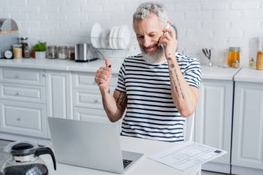Dövmeli adam, mutfaktaki bilgisayarın yanında akıllı telefondan konuşuyormuş gibi görünüyor. 