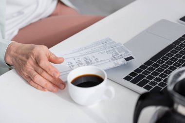 Kahve ve dizüstü bilgisayarın yanında faturaları tutan olgun bir kadın görüntüsü. 