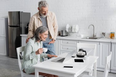 Mutfakta kahve ve aletlerle gülümseyen karısının yanında duran adam. 