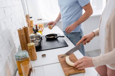 Cropped view of man seasoning pancake in frying pan near wife cutting banana in kitchen  clipart