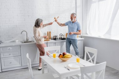 Gülümseyen olgun bir çiftin mutfakta spatulalarla kavga edişinin yan görüntüsü 