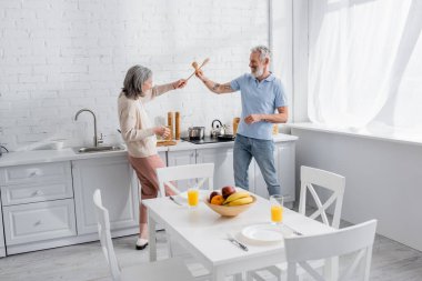 Side view of smiling mature couple fighting with spatulas in kitchen  clipart