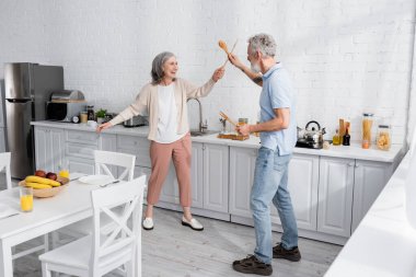 Happy mature woman fighting with wooden spatulas in kitchen  clipart