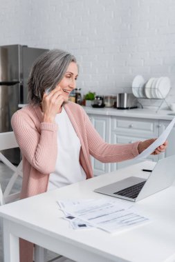 Gülümseyen olgun bir kadın akıllı telefonla konuşuyor ve mutfakta dizüstü bilgisayarının yanında kağıt tutuyor. 
