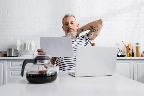 Starší Muž Drží Papíry Blízkosti Notebooku Rozmazané Konvice Kávy Kuchyni — Stock fotografie
