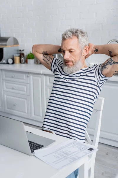 Татуированный Мужчина Смотрит Ноутбук Рядом Бумагами Счетами Кухне — стоковое фото