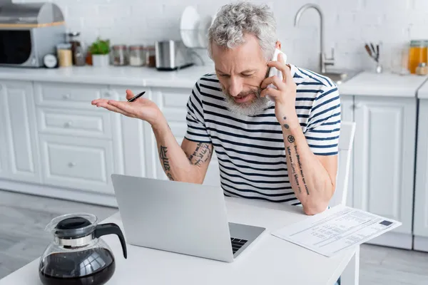 Yetişkin bir tele-işçi mutfakta bilgisayar ve kağıtların yanında akıllı telefondan konuşuyor. 