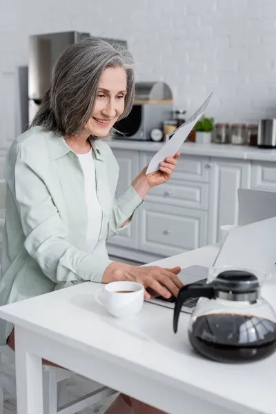 微笑着拿着帐单 在厨房边喝咖啡边使用笔记本电脑的女人 — 图库照片