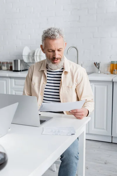 Gammal Man Tittar Papper Nära Laptop Köket Hemma — Stockfoto