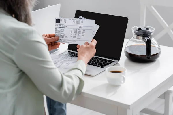 照片中 一个成熟的女人拿着钞票 靠近笔记本电脑 在厨房里有空白屏幕和咖啡壶 — 图库照片