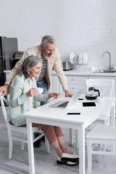 一个成熟的女人指着靠近丈夫的笔记本电脑和厨房里的咖啡 — 图库照片