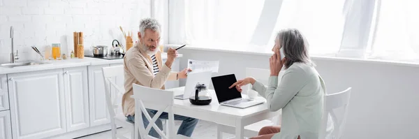 Hombre Apuntando Esposa Hablando Teléfono Inteligente Cerca Facturas Portátiles Cocina —  Fotos de Stock