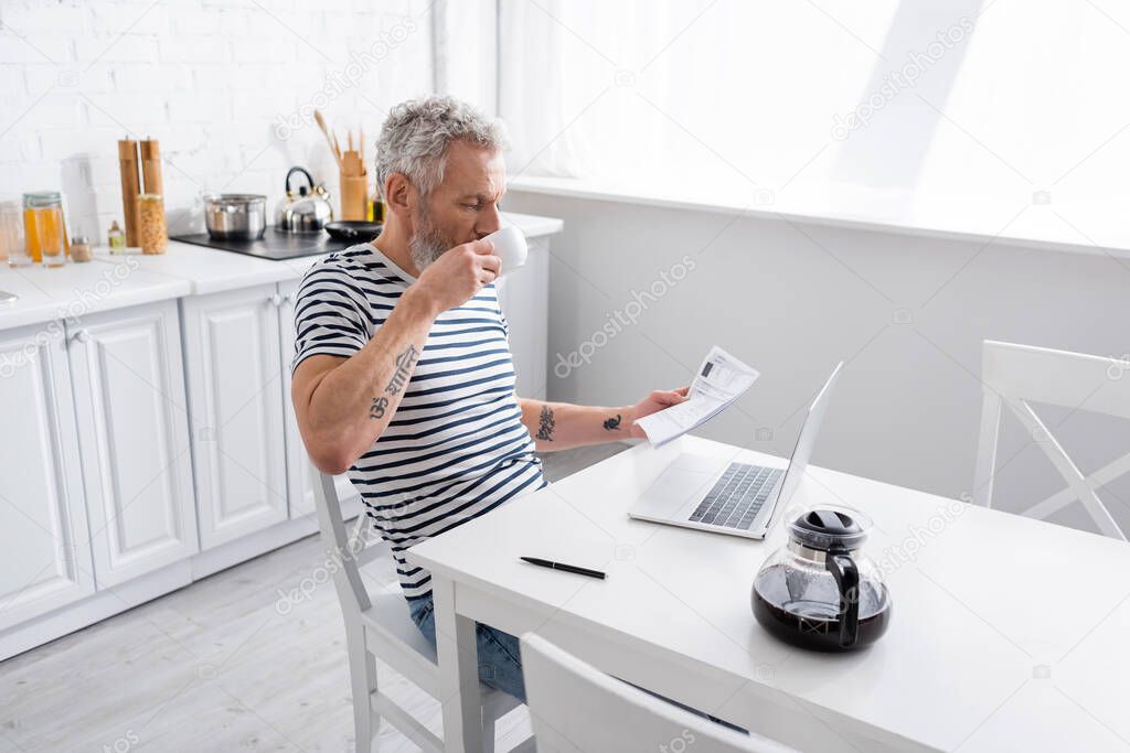 man holding papers with bills and drinking coffee near laptop in kitchen. Translation: 